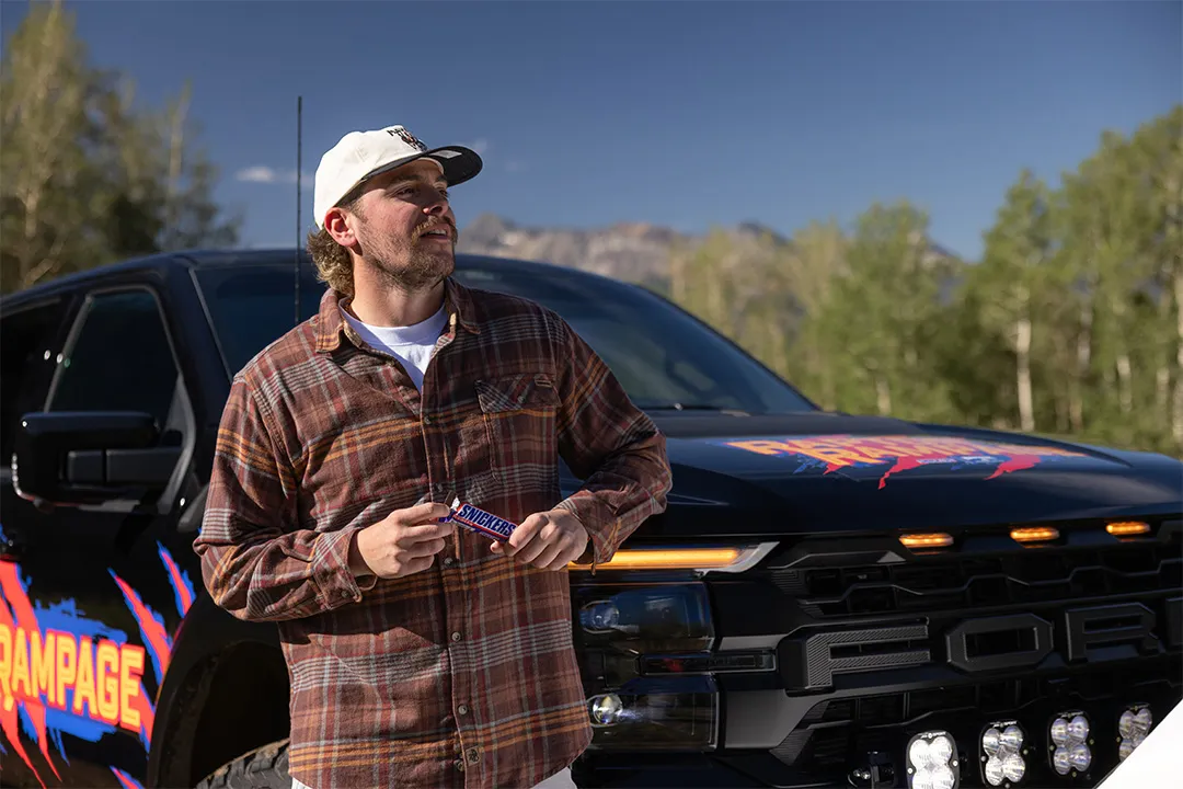 Man standing next to Raptor Rampage prize vehicle eating a SNICKERS® candy bar