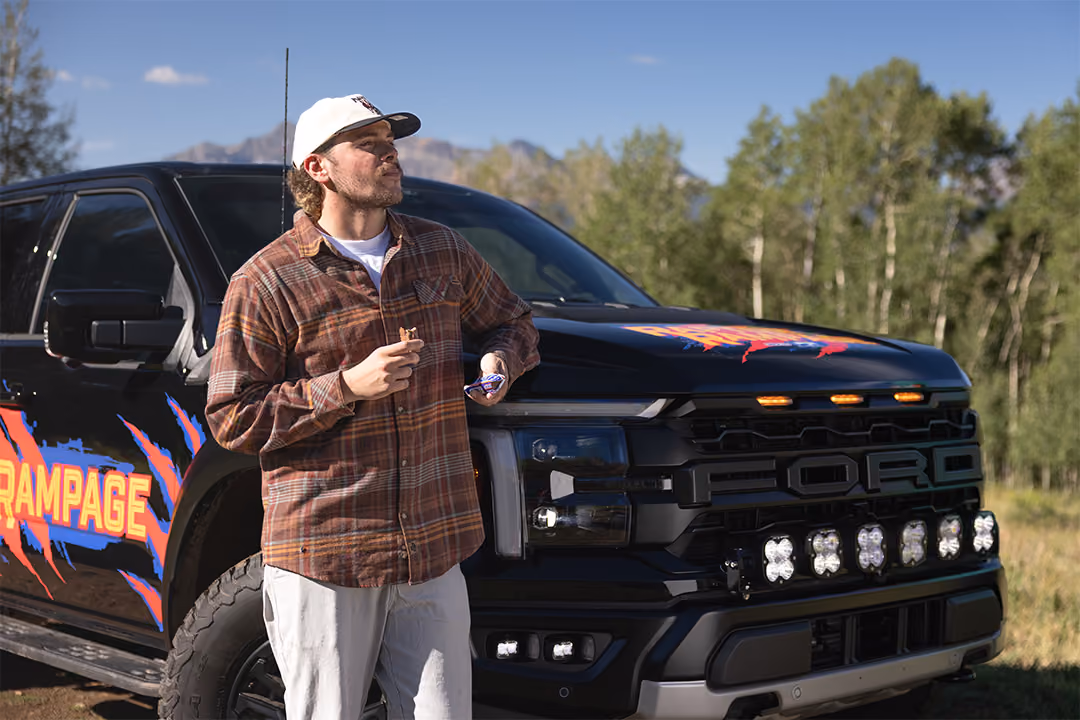 Man standing next to Raptor Rampage prize vehicle eating a SNICKERS® candy bar